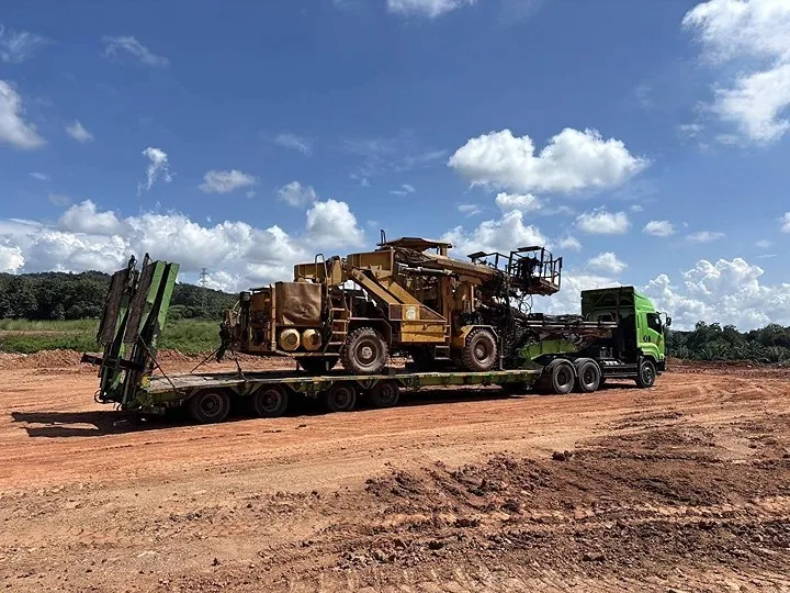 Heavy mining excavator loaded on a lowboy trailer being hauled by a green semi-truck on a dirt site under a blue sky